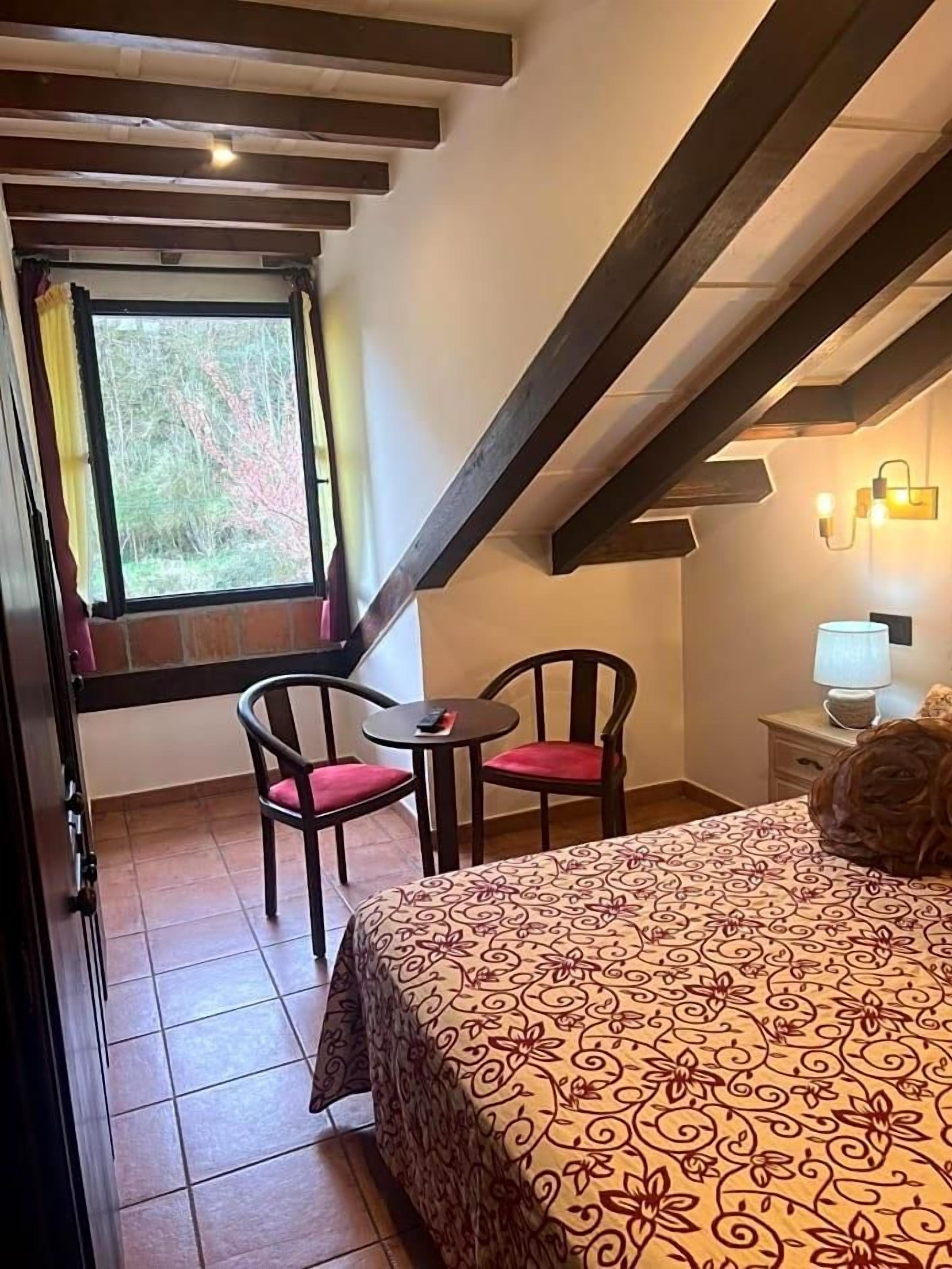 Un dormitorio con cama, dos sillas, una mesita, una ventana con cortinas y una lámpara en una mesita de noche.