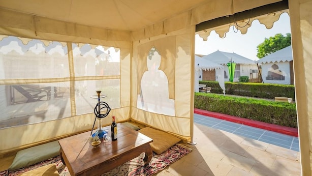 Una habitación con tienda de campaña, una mesa de madera, un narguile y vistas a un jardín y edificios.