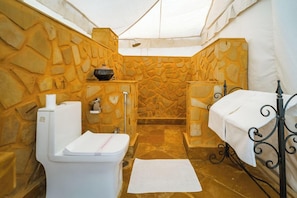 A compact bathroom with a stone wall, a toilet, a sink, and a small table with a towel. - The Mamas Resort and Camp (Jaisalmer)