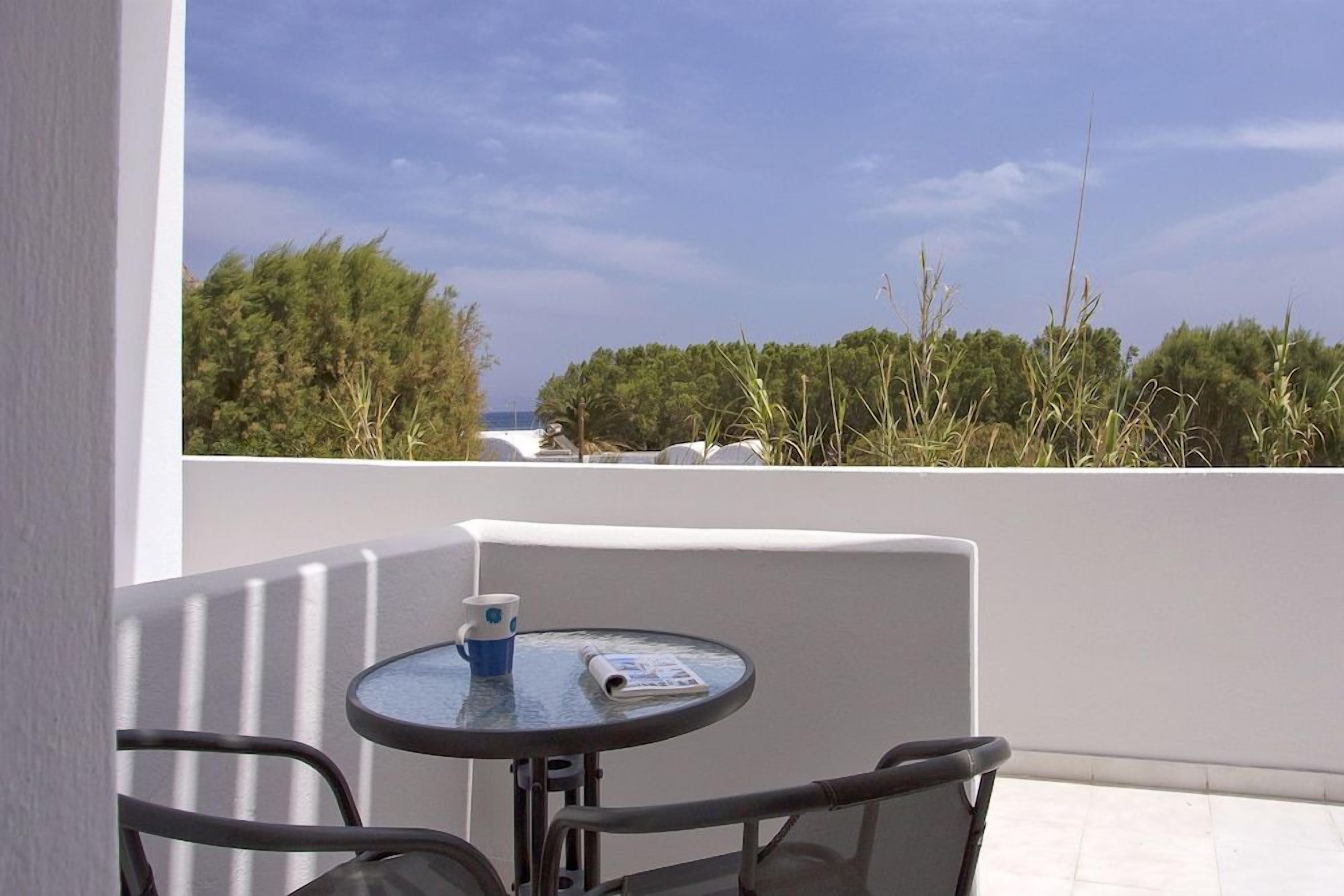 Ein Balkon mit Tisch und Stühlen, der auf eine Küstenlandschaft mit Vegetation und blauem Himmel blickt.