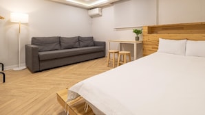 Ein Hotelzimmer mit einem Bett, einem grauen Sofa, einem kleinen Tisch mit zwei Hocker und einer Stehlampe.