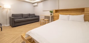 Una habitación de hotel con una cama, un sofá gris, una pequeña mesa con dos taburetes y una lámpara de pie.