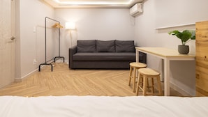 Ein gemütlicher Raum mit Bett, grauem Sofa, kleinem Tisch mit Hocker, Pendelleuchte und einem Blumentopf.