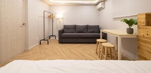Una habitación funcional con cama, sofá gris, mesita con taburetes, lámpara colgante y planta en maceta.