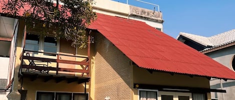 A two-story building with a red roof, a balcony, and a window with curtains.