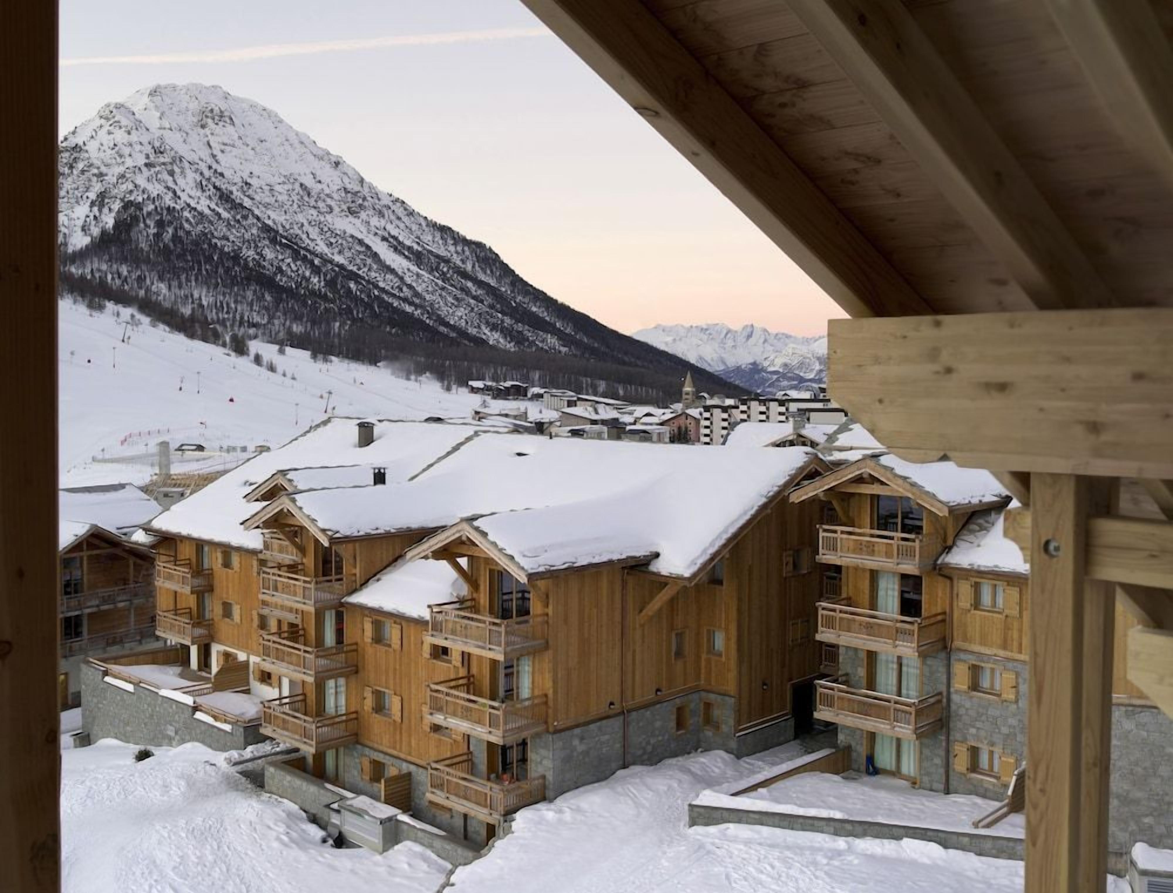 Un edificio in stile chalet in legno con balconi, circondato da montagne innevate e una località sciistica sullo sfondo.