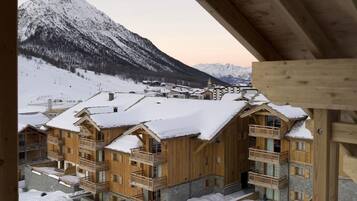 Un edificio in stile chalet in legno con balconi, circondato da montagne innevate e una località sciistica sullo sfondo.