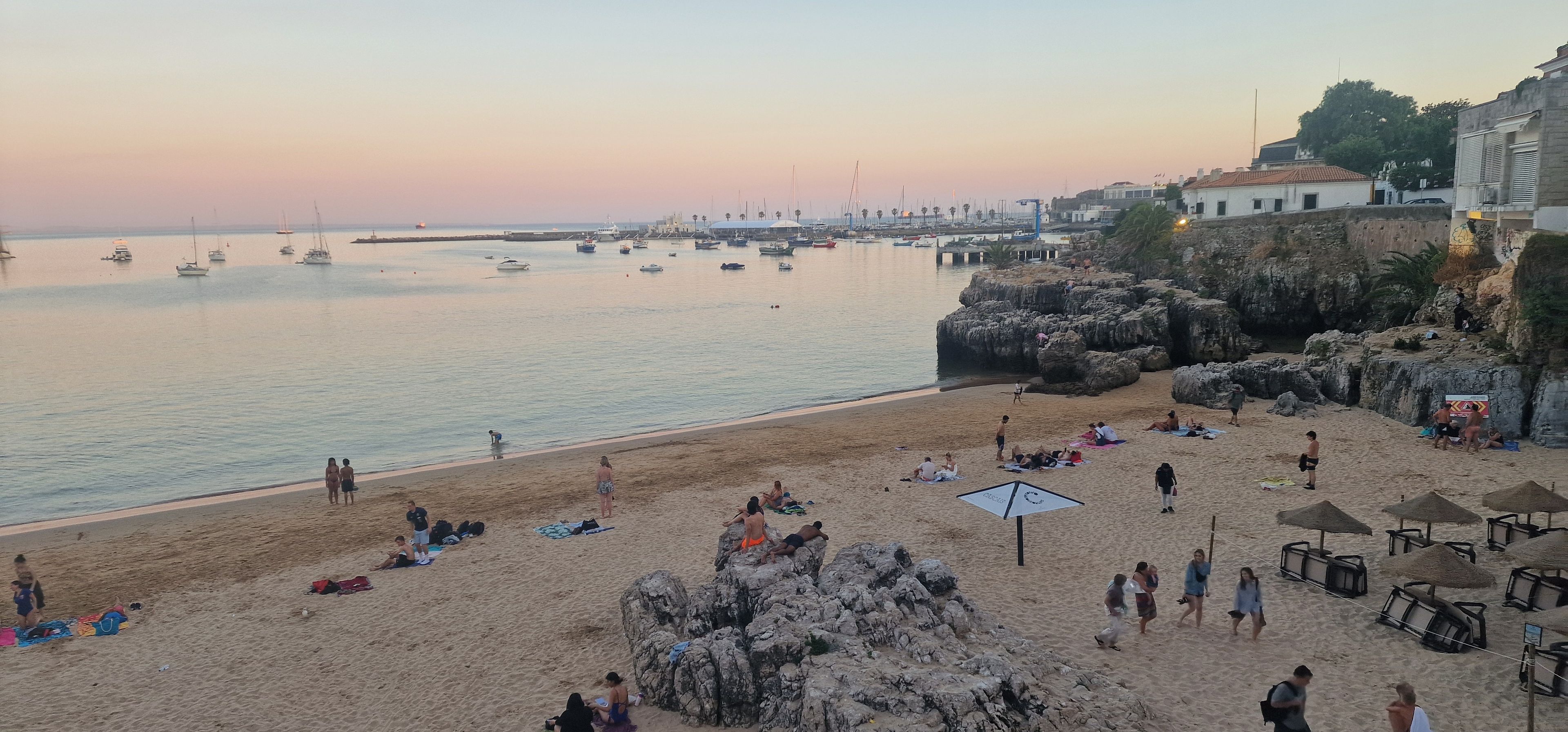 Ein Strand mit badenden Personen und Spaziergängern, einem Felsen und Booten im Wasser.