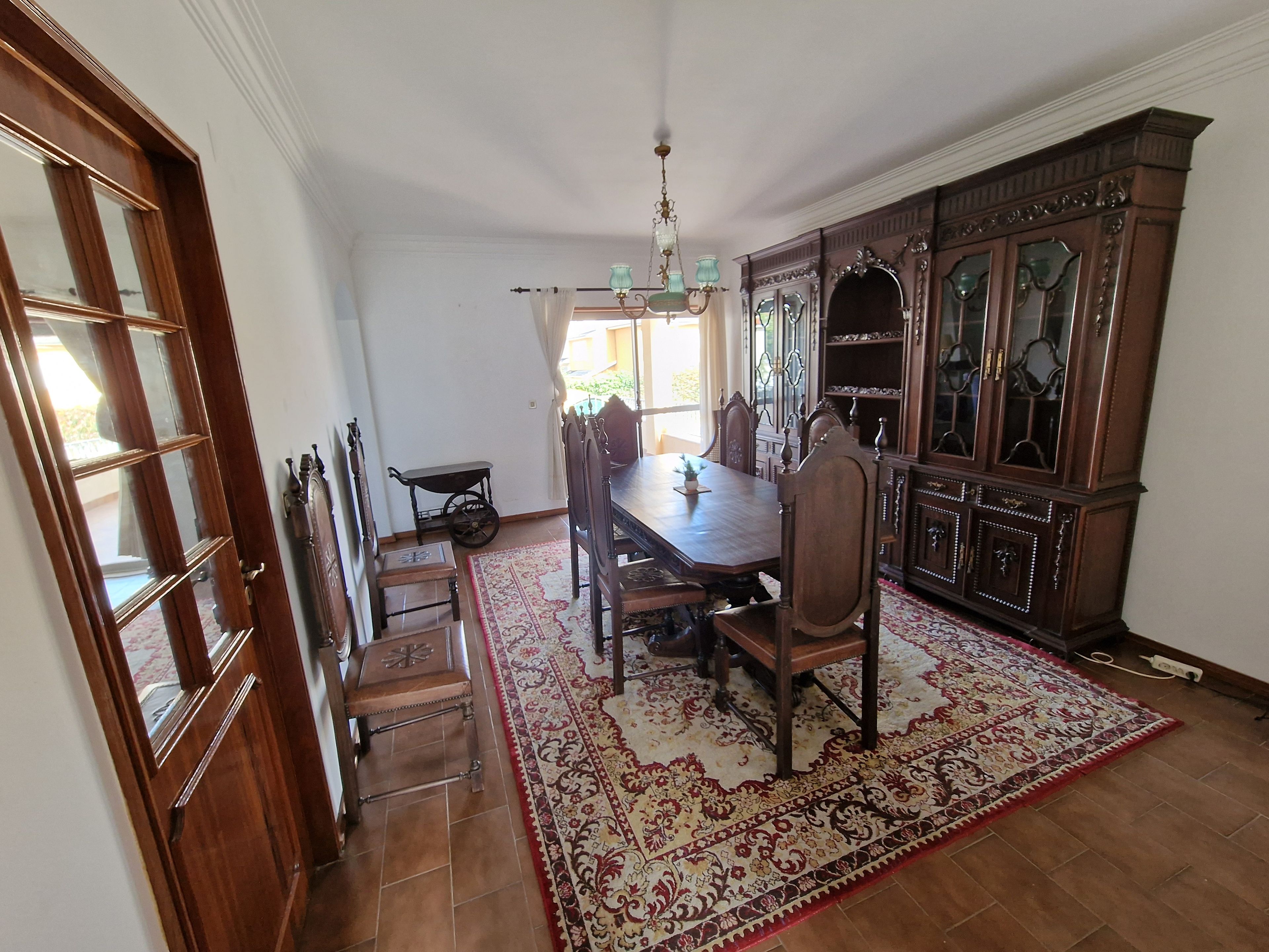 A dining room with a wooden table, chairs, a chandelier, and a patterned rug.