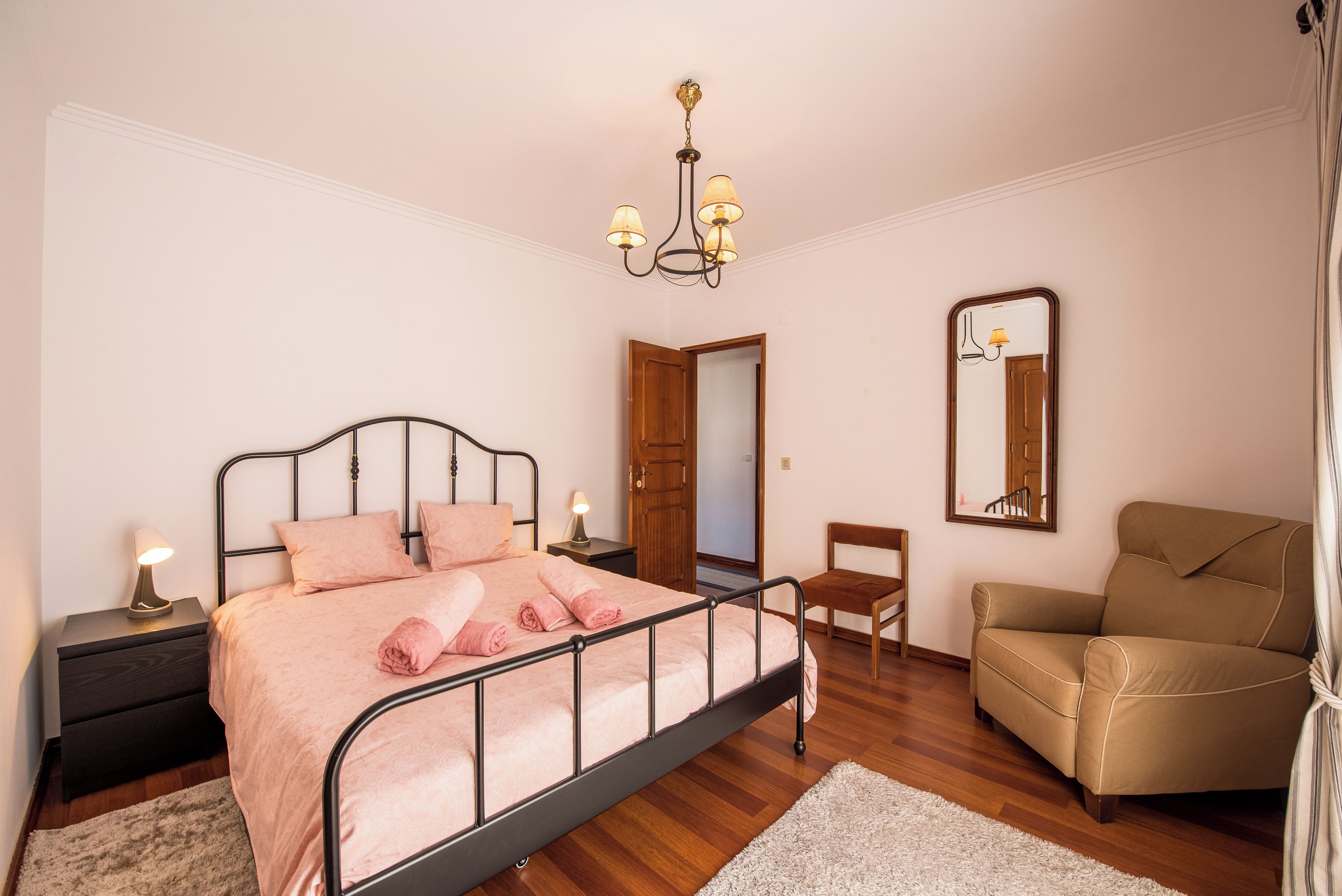 Ein Schlafzimmer mit einem Bett, Nachttischlampen, einer Holztür, einem Sessel, einem Spiegel und einer Chandelier.