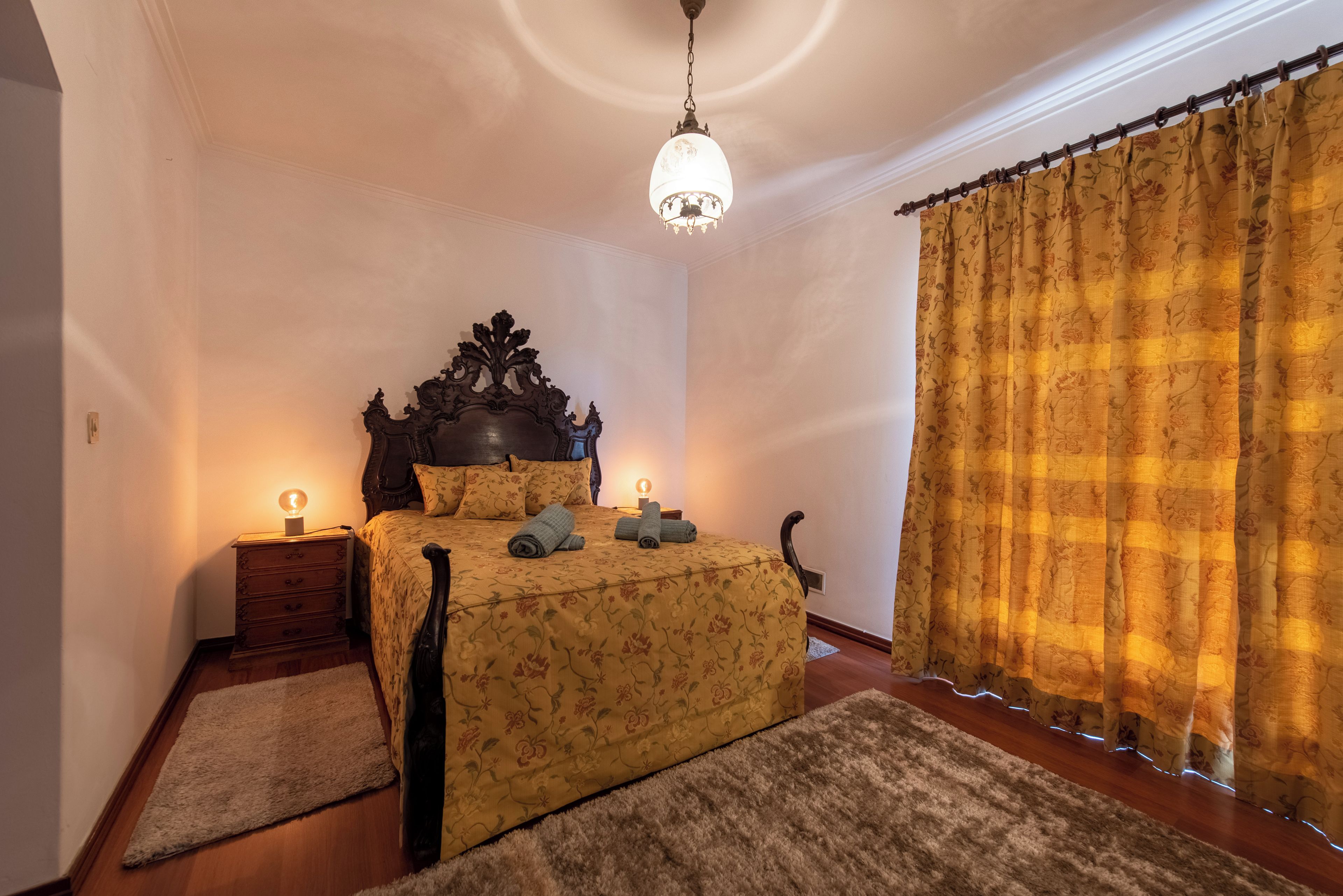 Ein Schlafzimmer mit einem hölzernen Bett, einer Kristallleuchter, einem Nachttisch und einem großen Fenster mit Vorhängen.