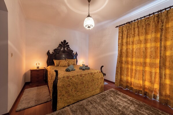 Ein Schlafzimmer mit einem hölzernen Bett, einer Kristallleuchter, einem Nachttisch und einem großen Fenster mit Vorhängen.