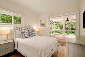 Ein Schlafzimmer mit einem Bett, Nachttischen, einer Lampe, einem gerahmten Bild und Blick auf einen Garten durch große Fenster.