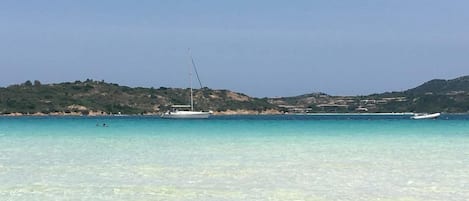 A coastal scene with clear turquoise water, boats, and a hilly landscape in the background.