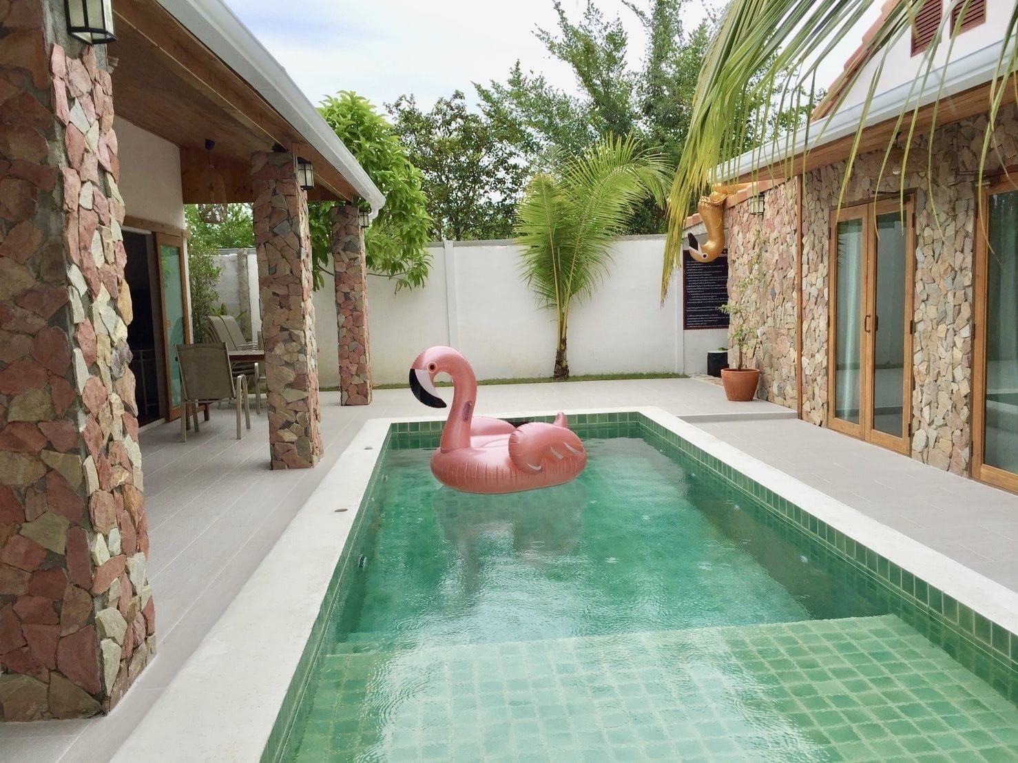 Una piscina con un flotador de flamenco rosa, paredes de piedra y una zona de patio cubierta.