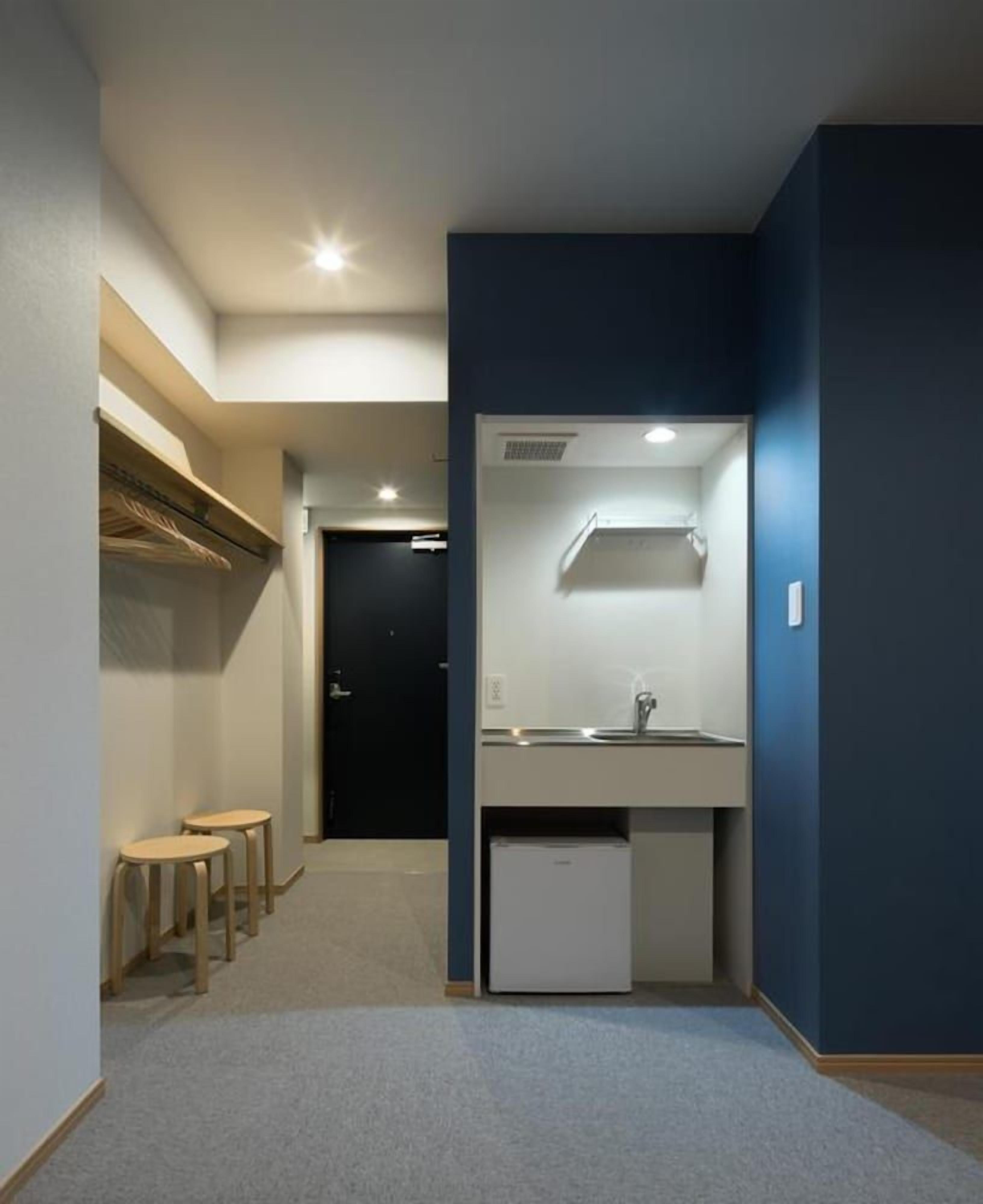 A narrow hallway with a small kitchenette area, including a sink and a refrigerator. There are two stools and a door leading to another room.