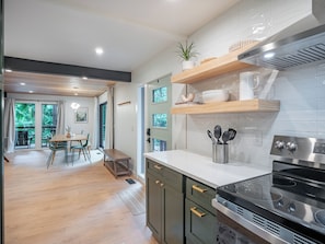 A modern kitchen with a built-in stove, wooden shelves, and a dining area with a table and chairs. - Riverfront Mt. Hood Retreat with Hot Tub + Game Room (Rhododendron)
