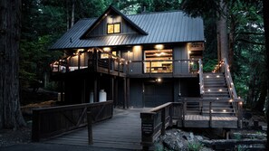 A two-story wooden cabin with a deck and stairs, surrounded by forest. - Riverfront Mt. Hood Retreat with Hot Tub + Game Room (Rhododendron)