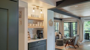 A modern kitchen with a coffee machine, shelves with white mugs, and a dining area with a table and chairs. - Riverfront Mt. Hood Retreat with Hot Tub + Game Room (Rhododendron)