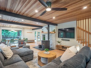 A modern living room with a fireplace, a ceiling fan, and a large TV. - Riverfront Mt. Hood Retreat with Hot Tub + Game Room (Rhododendron)