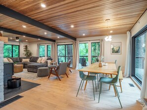 A modern living room with a sectional sofa, a dining table with chairs, and large windows offering a view of the outdoors. - Riverfront Mt. Hood Retreat with Hot Tub + Game Room (Rhododendron)
