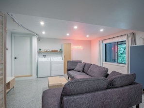 A modern living room with a grey sofa, a washing machine, and a window with a view of greenery. - Riverfront Mt. Hood Retreat with Hot Tub + Game Room (Rhododendron)