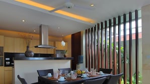 A modern kitchen with a dining area, featuring a central island, dark wicker chairs, and a view through a glass partition.
