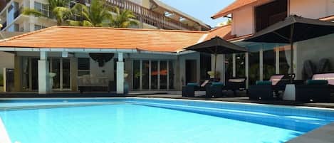 A swimming pool with lounge chairs and umbrellas, a covered seating area, and a building with a tiled roof.