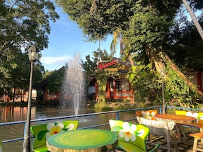 Un restaurante junto al río con terraza, una fuente y un letrero con escritura tailandesa.