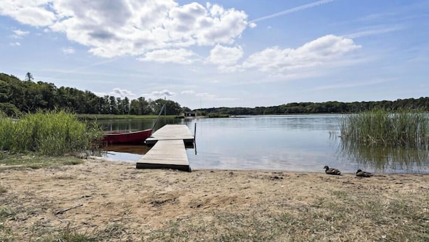 Un lago tranquilo con un muelle de madera que se adentra en el agua, rodeado de orillas cubiertas de hierba y árboles.