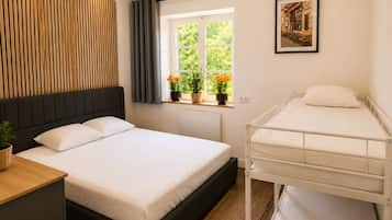 Ein Hotelzimmer mit Bett, Nachttisch, einem Fenster mit Vorhängen, einer Heizung, einem Wandregal mit Pflanzen und einem gerahmten Bild an der Wand.