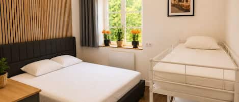 A hotel room with a bed, a bedside table, a window with curtains, a radiator, a wall-mounted shelf with plants, and a framed picture on the wall.