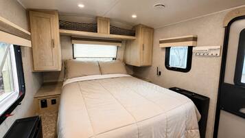 A compact RV interior with a bed, built-in cabinets, and a small window.