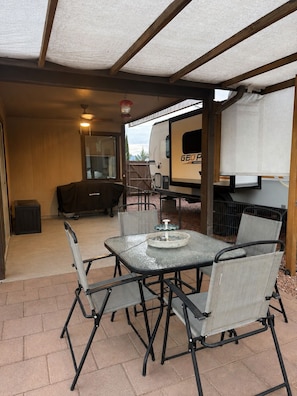 Une terrasse extérieure avec une table et des chaises, un barbecue et un espace abrité où est garé un camping-car à proximité.