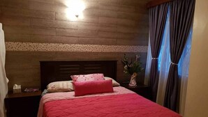 A bedroom with a wooden headboard, a red bedspread, a nightstand with a lamp, and a window with curtains. - Alkasim (Nairobi)