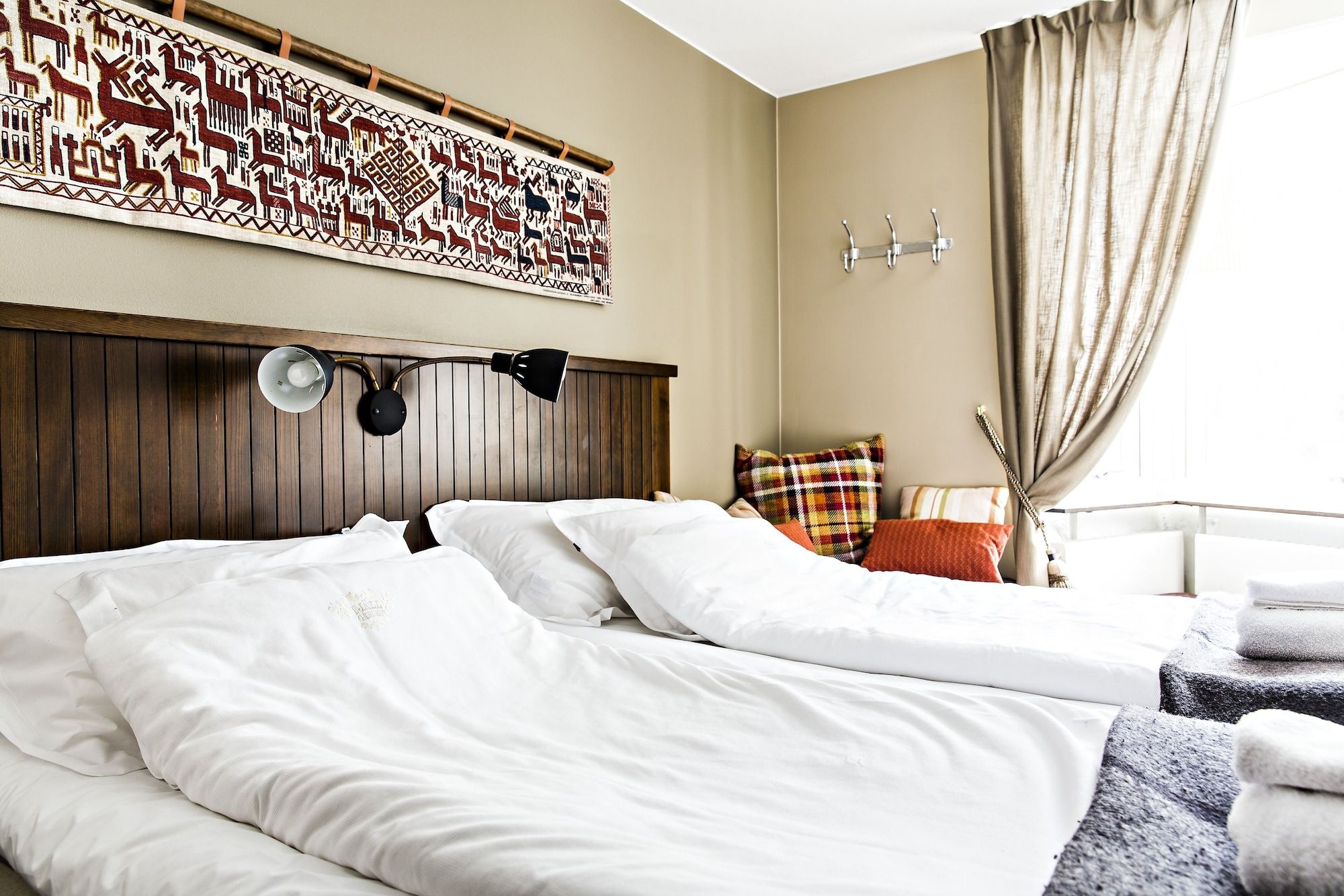 A hotel room with a wooden headboard, a bed with white linens, a decorative wall hanging, a bedside lamp, and a window with curtains.