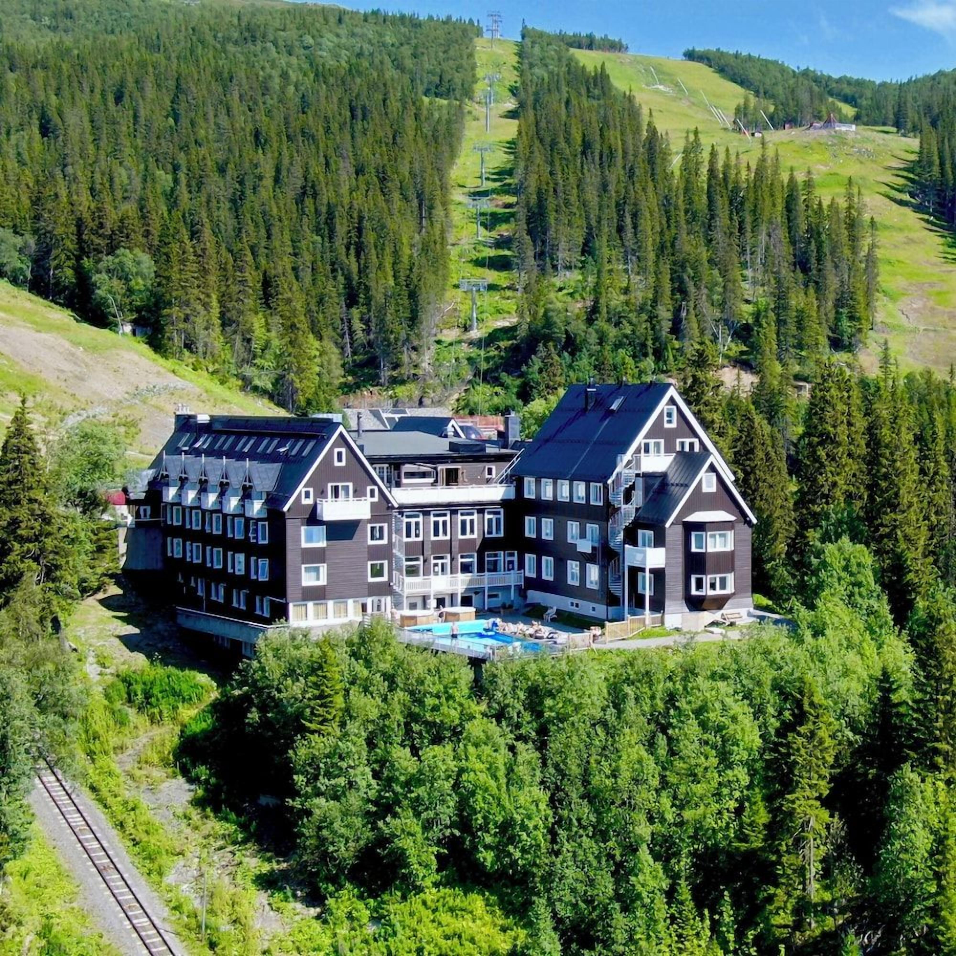 A multi-story hotel with a swimming pool, surrounded by dense forest and hills.