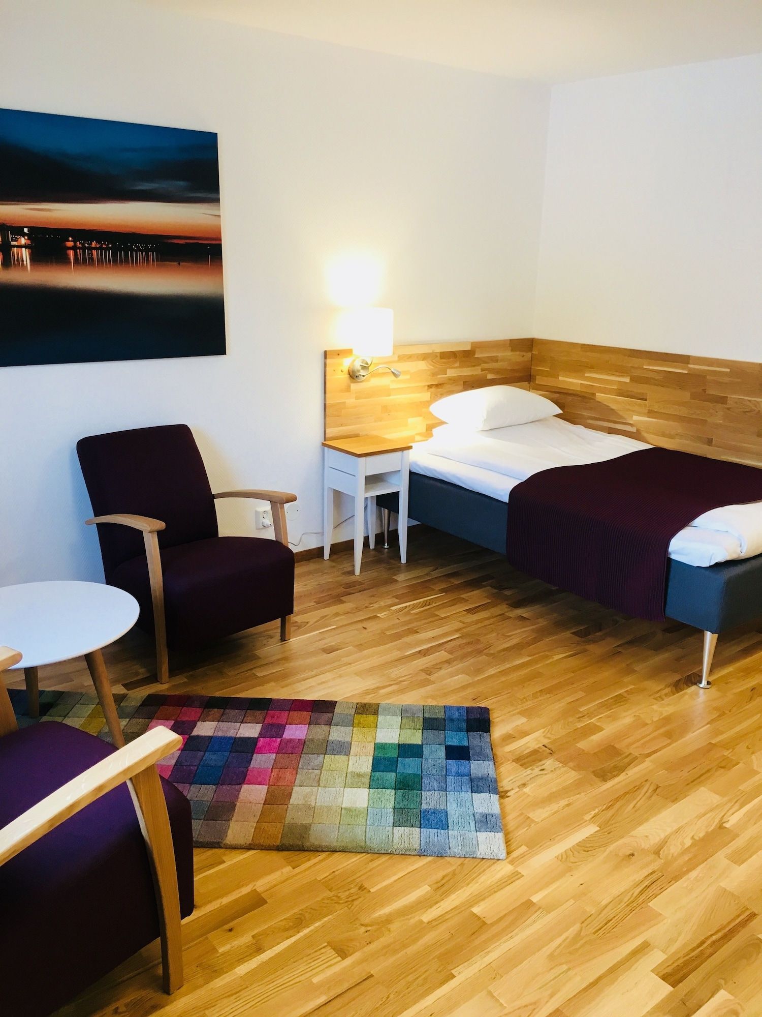 Ett hotellrum med en säng, en stol, ett litet bord, en lampa och en färgglad matta.