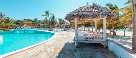 A swimming pool with a stone deck, a thatched-roof gazebo, and palm trees.