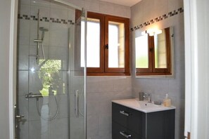 Un baño con ducha envuelta en vidrio, tocador con espejo y ventana sobre el lavabo.