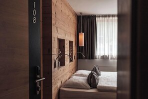 Hotellihuone, jossa on puinen seinä, tyynyillä varustettu sänky, riippuvalaisin ja ikkuna, jossa on verhot.