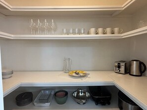 Una cocina con estantes blancos, un tostador, una tetera y diversos utensilios de cocina.