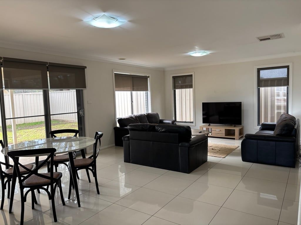 A spacious living room with a glass dining table, two sofas, a flat-screen TV, and large windows.