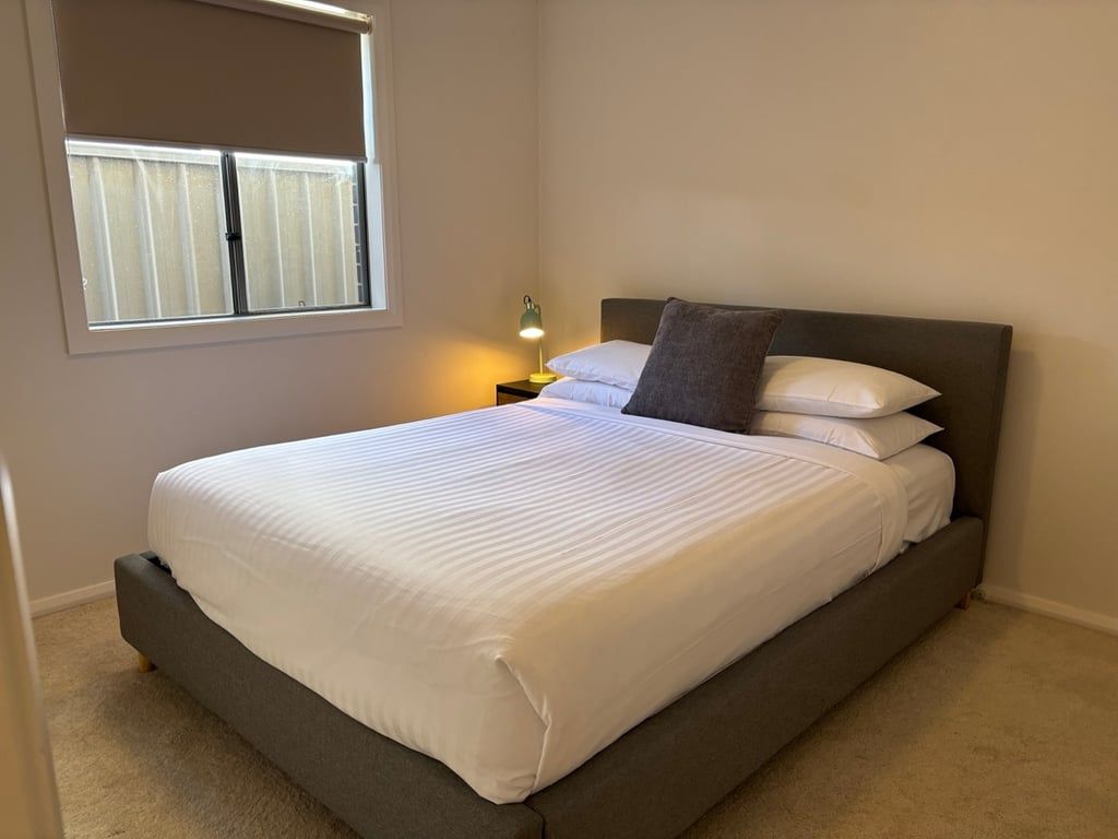 A neatly made bed with white linens in a room with a window and a nightstand with a lamp.