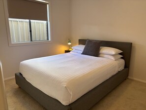 A neatly made bed with white linens in a room with a window and a nightstand with a lamp.