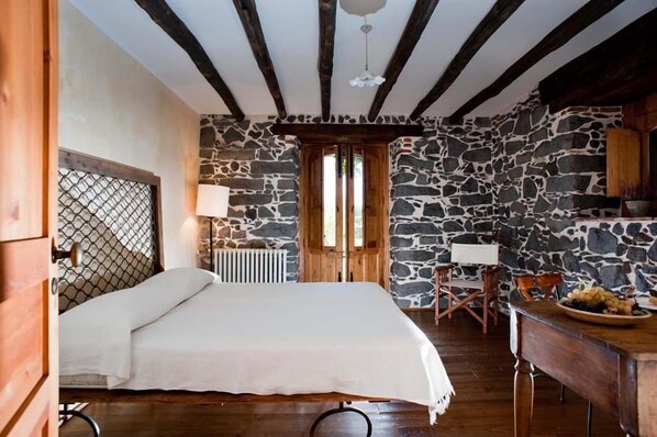 Un dormitorio con pared de piedra, vigas de madera en el techo, una cama, un radiador y una zona de comedor con mesa y sillas.
