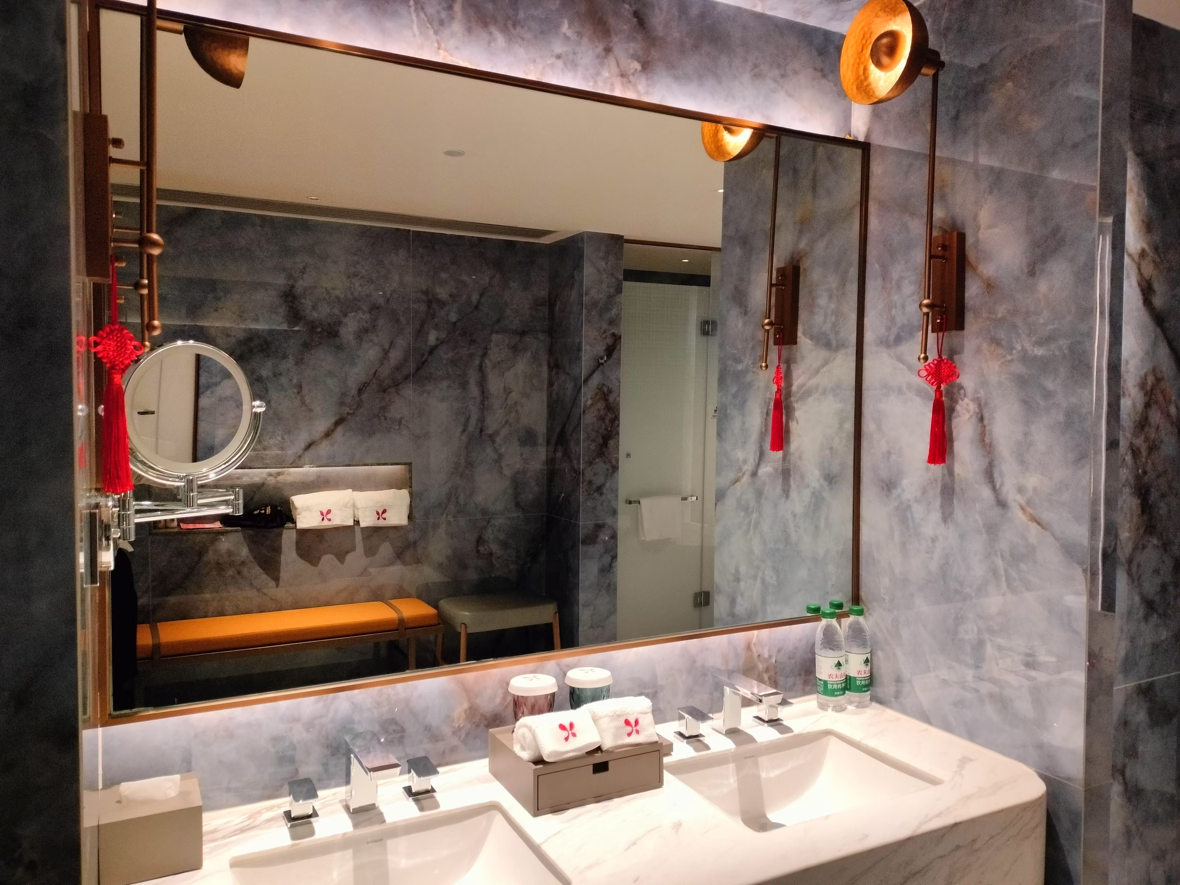 A modern bathroom with a large mirror, marble walls, a sink with multiple faucets, and a bench in the background.