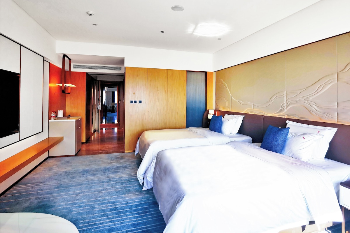 Quarto de hotel com duas camas, uma escrivaninha, televisão e banheiro visível por uma porta aberta.