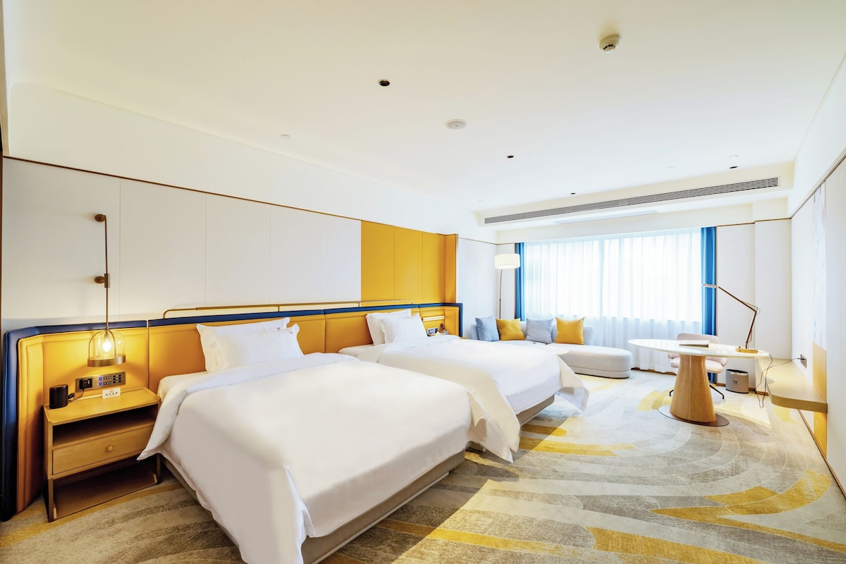 Quarto de hotel com duas camas, uma escrivaninha, uma cadeira e uma janela grande com cortinas.