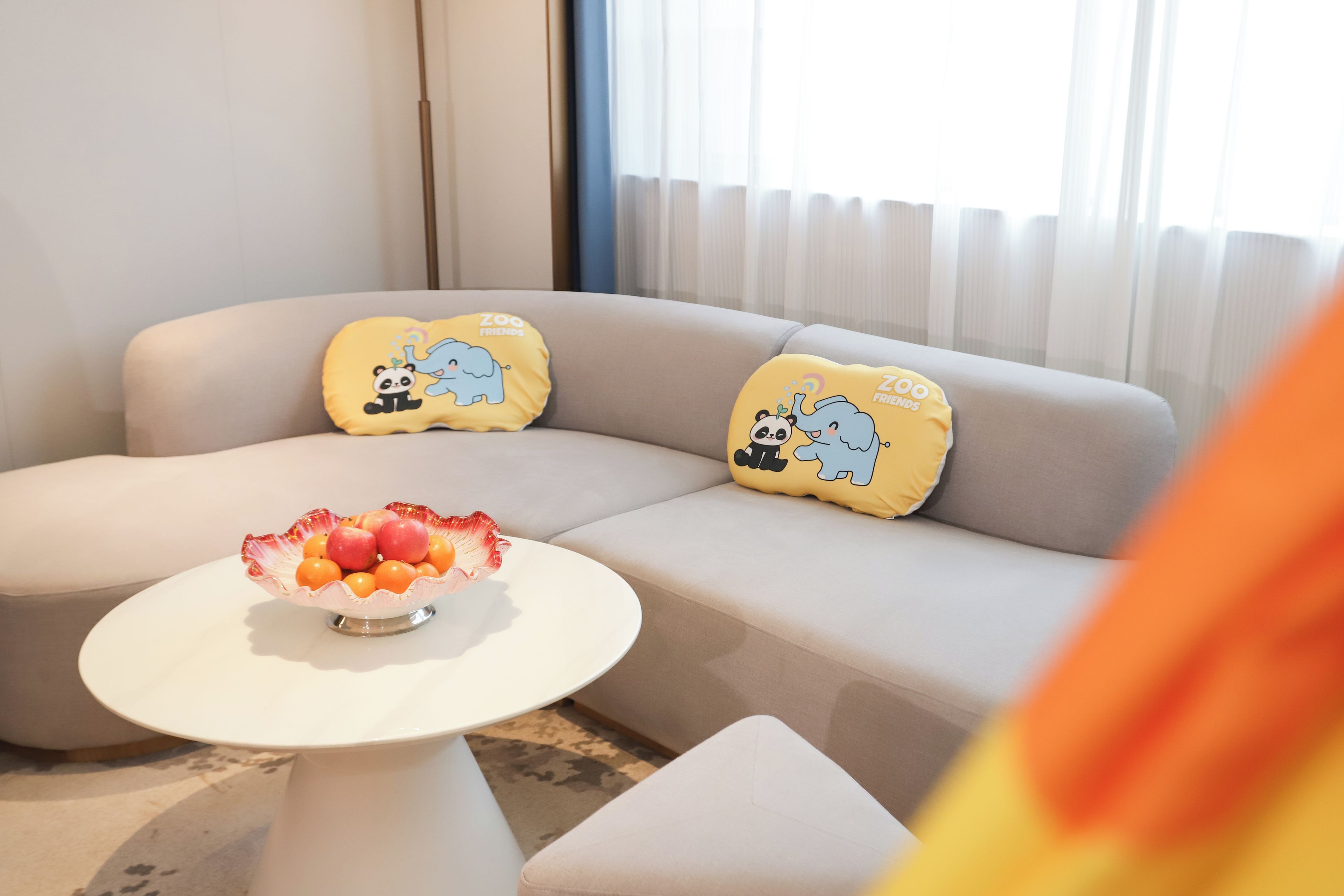 A modern living room with a beige sofa, two yellow pillows with cartoon animals, and a white coffee table with a fruit bowl.